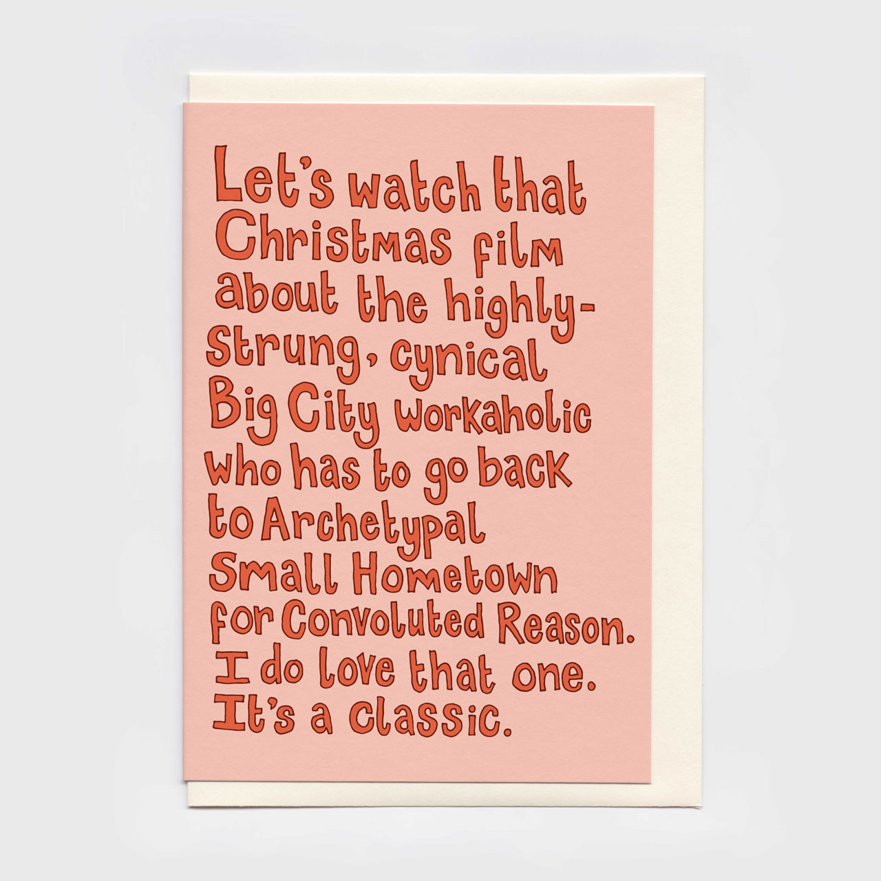A6 Christmas card with a light pink front and a cream envelope behind it, featuring a humorous quote about a Christmas film in red, hand-drawn blocky lettering.