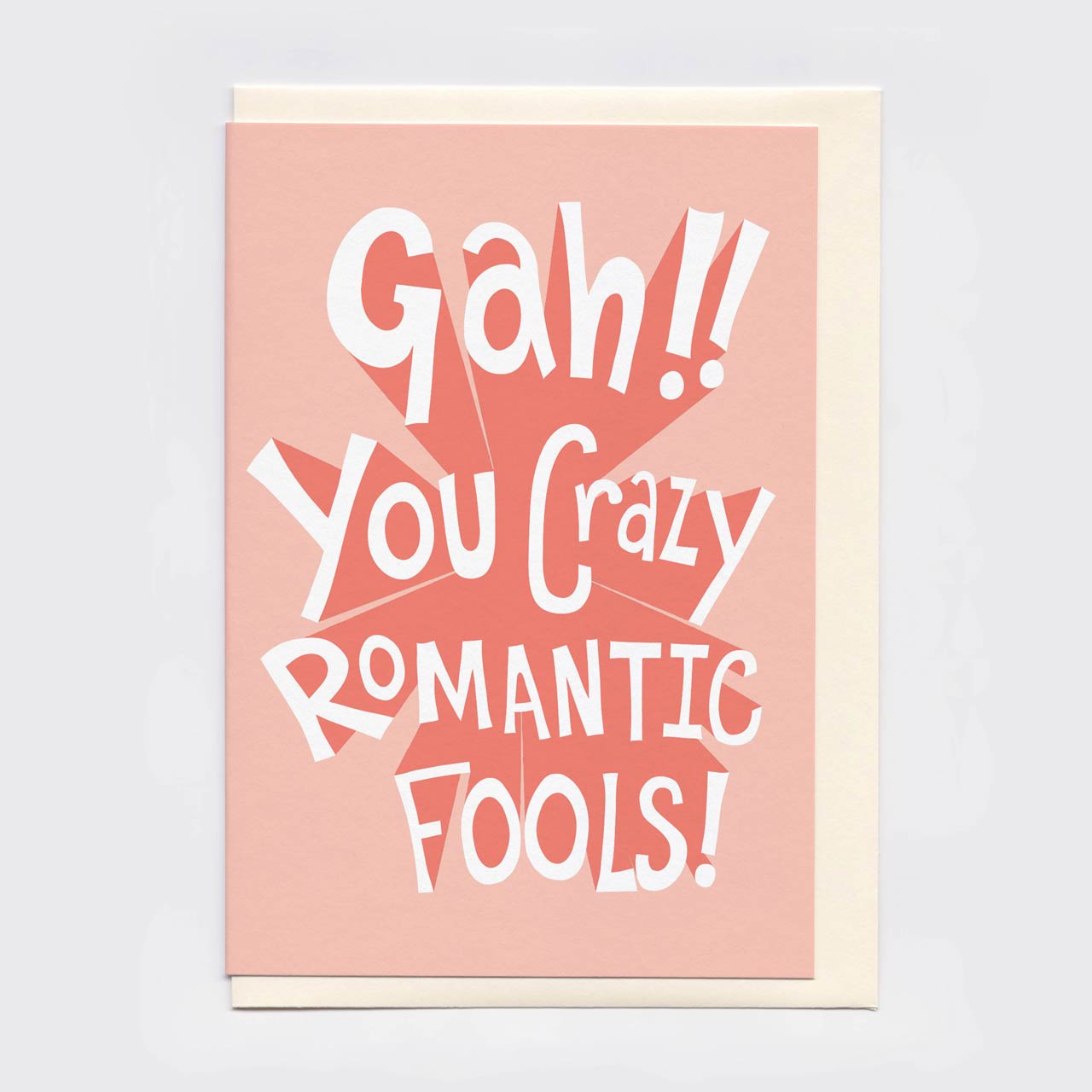 An A6 greeting card with a light pink front and cream envelope, featuring the phrase 'Gah!! You Crazy ROMANTIC FOOLS!' in large white retro lettering over a red burst graphic, shown on a light grey background.