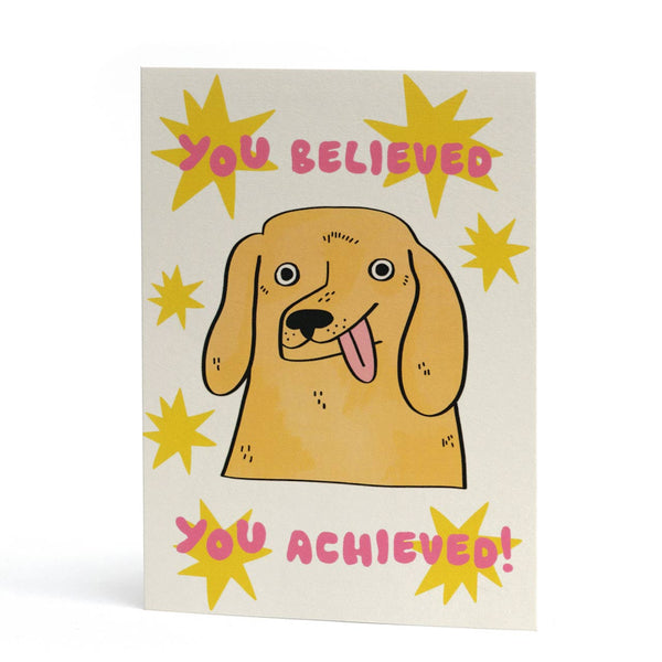 A funny congrats card featuring a stylised illustration of a golden retriever and the words "You believed, you achieved!" in pink.