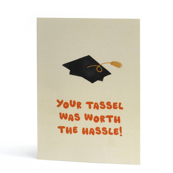 A funny rhyming graduation card with a black mortarboard cap and gold tassel illustration above orange text that reads, 'YOUR TASSEL WAS WORTH THE HASSLE!'