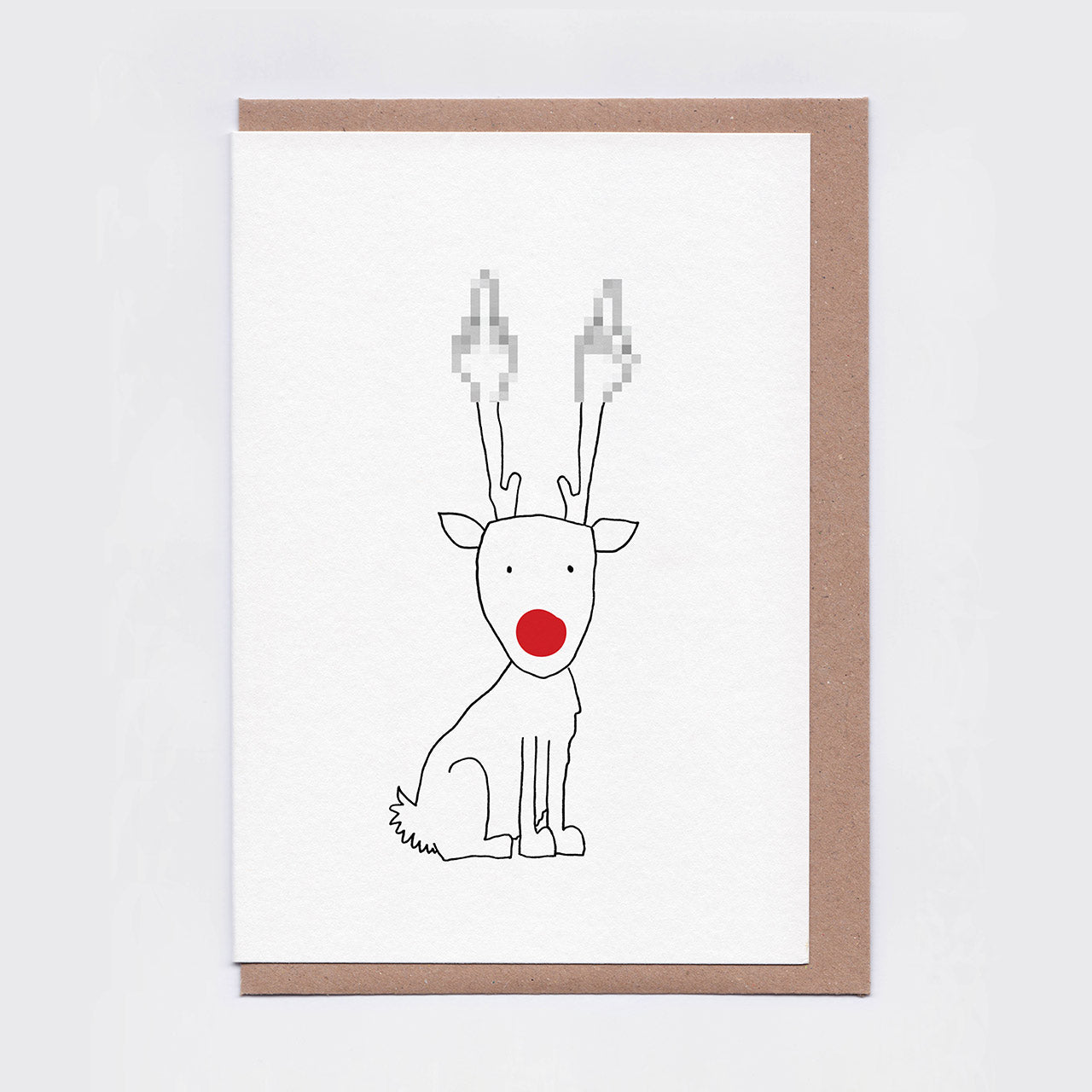 A white greeting card featuring a minimalist black line drawing of a reindeer with a bright red nose and pixellated antlers, shown with a brown kraft envelope behind it on a light grey background.