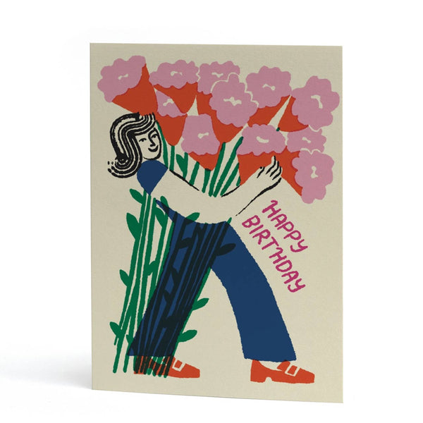 An A6 greeting card featuring a retro-style illustration of a woman holding a large bouquet of pink flowers, with the words 'HAPPY BIRTHDAY' printed in pink along the side. The card has a warm cream background and is photographed standing upright against a clean white background.