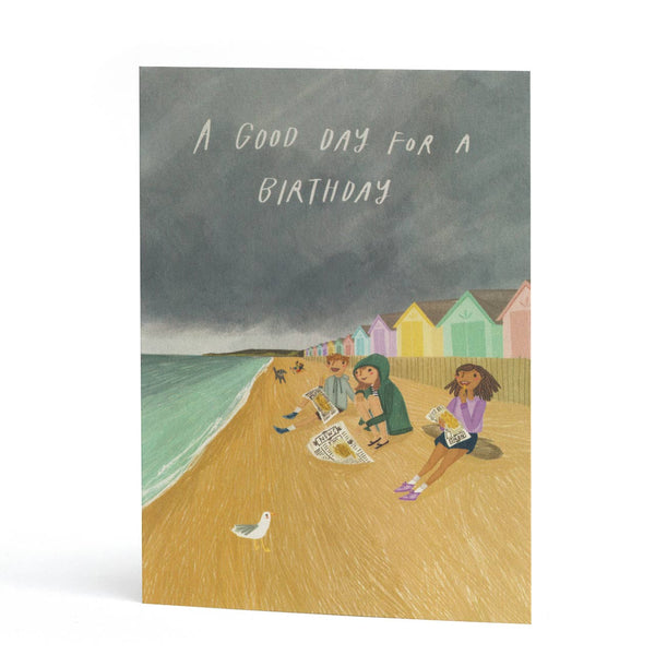 A greeting card featuring an illustration of children on a beach with seagulls and pastel beach huts under a dark gray sky. The text at the top reads 'A GOOD DAY FOR A BIRTHDAY'.