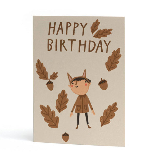 An A6 greeting card with a warm beige background, featuring the words 'HAPPY BIRTHDAY' in brown lettering, surrounded by illustrated brown oak leaves, acorns, and a cute cartoon child in a brown animal-ears outfit.