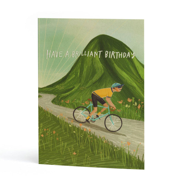 A vertical greeting card featuring an illustration of a cyclist riding along a winding road in front of a large green mountain. The text 'HAVE A BRILLIANT BIRTHDAY' is printed across the top.