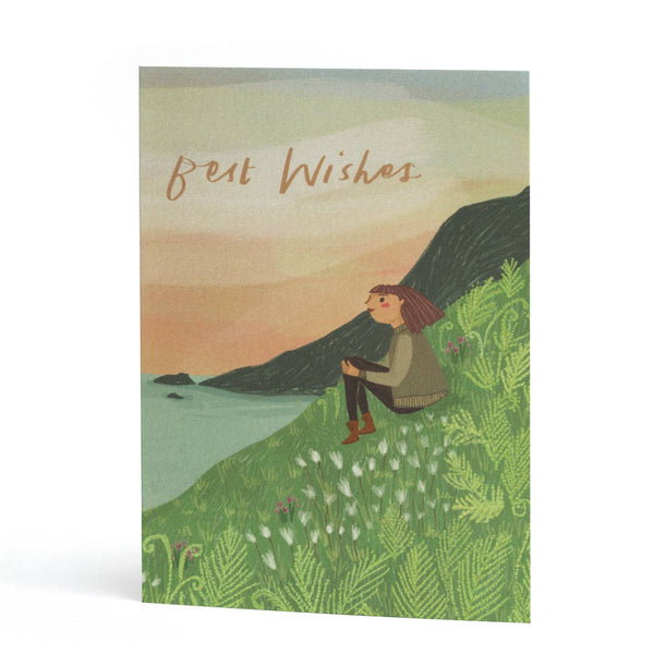 A greeting card featuring an illustration of a woman sitting on a grassy hill overlooking water, with the words 'Best Wishes' written in brown script at the top.