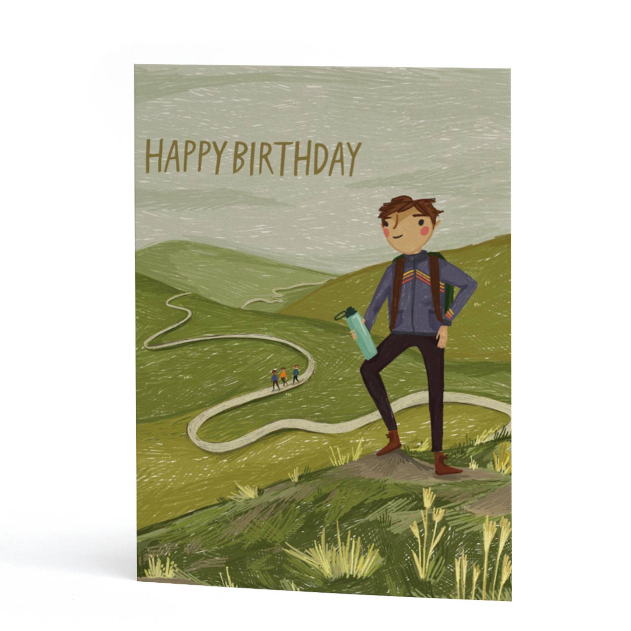 An A6 greeting card featuring a hand-drawn illustration of a hiker standing on a grassy hill with rolling green hills and a winding path in the background. The text 'HAPPY BIRTHDAY' is printed at the top.