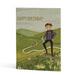 An A6 greeting card featuring a hand-drawn illustration of a hiker standing on a grassy hill with rolling green hills and a winding path in the background. The text 'HAPPY BIRTHDAY' is printed at the top.