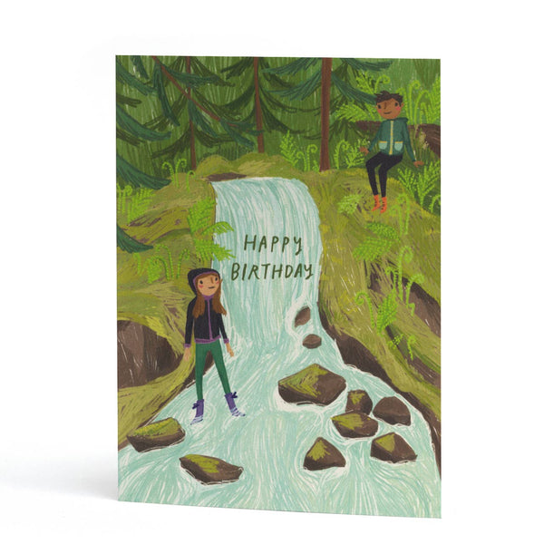 An A6 portrait birthday card featuring a hand-drawn illustration of a forest waterfall with two people, one standing in the stream and one sitting on a rock. The text 'Happy Birthday' is written across the center.