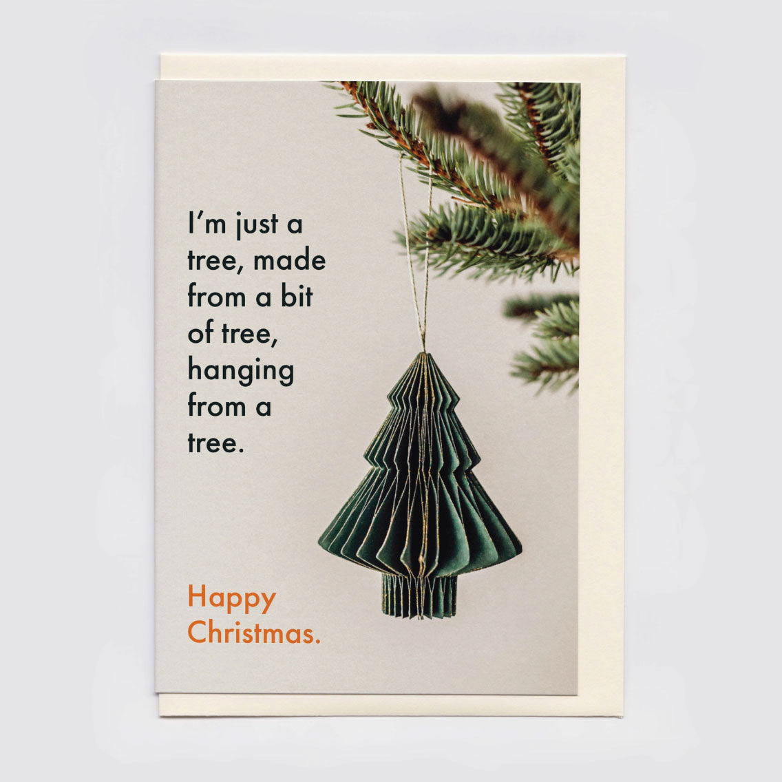 A Christmas greeting card sitting on a cream envelope, featuring a green hanging tree ornament, pine branches at the top right, and the text, 'I'm just a tree, made from a bit of tree, hanging from a tree. Happy Christmas.