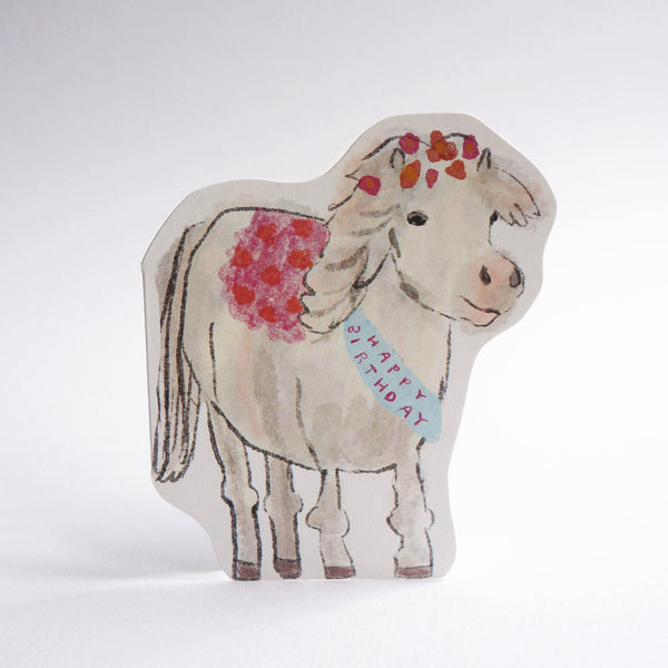 A die-cut greeting card featuring a hand-drawn illustration of a cream coloured pony with red and pink floral accents on its mane and back, wearing a blue 'HAPPY BIRTHDAY' ribbon, set against a plain light grey background.