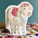 A handmade paper cutout of a white pony decorated with red floral patterns, a blue 'HAPPY BIRTHDAY' ribbon, and a red flower crown, displayed on a floral envelope, set against a blue background.