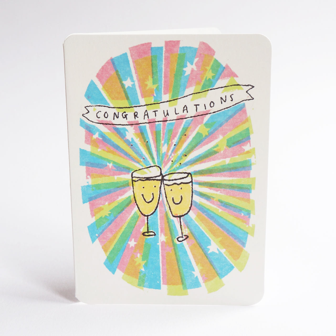 An A6 congratulations card with rounded corners, featuring a colourful burst design in pastel pink, blue, green, and yellow with two clinking champagne glasses and the word 'CONGRATULATIONS' on a banner, shown against a white background.