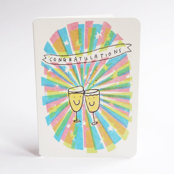 An A6 congratulations card with rounded corners, featuring a colourful burst design in pastel pink, blue, green, and yellow with two clinking champagne glasses and the word 'CONGRATULATIONS' on a banner, shown against a white background.