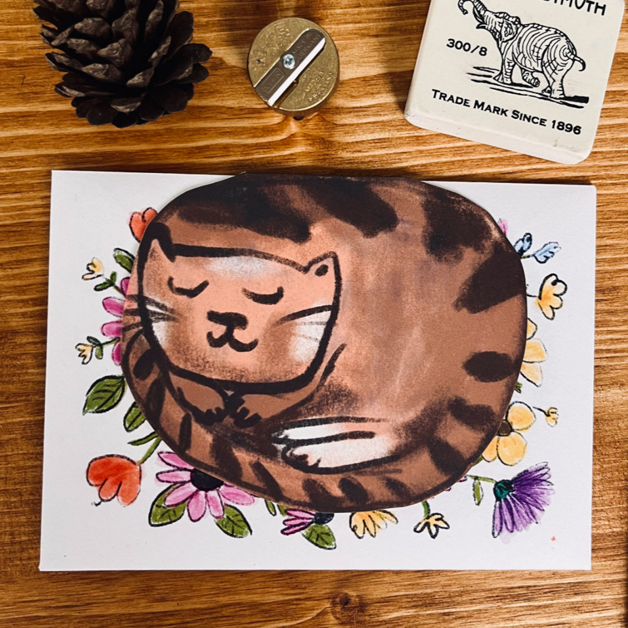 A die-cut card featuring a sleeping brown tabby cat, resting on a floral envelope, placed on a wooden surface with a pinecone, a pencil sharpener, and a small eraser nearby.