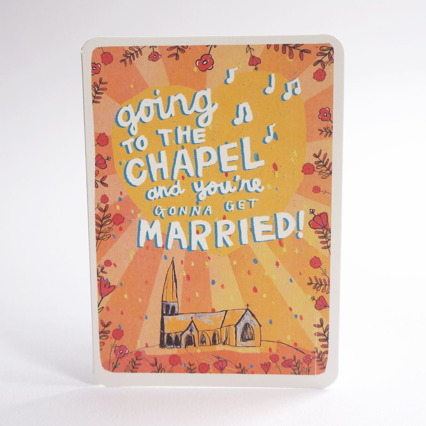 An A6 greeting card with rounded corners, featuring the phrase “going to the CHAPEL and you’re gonna get MARRIED!” in large blue and white lettering over a warm orange and yellow sunburst background, decorated with floral motifs and a small church illustration. The item is photographed alone on a clean white background.