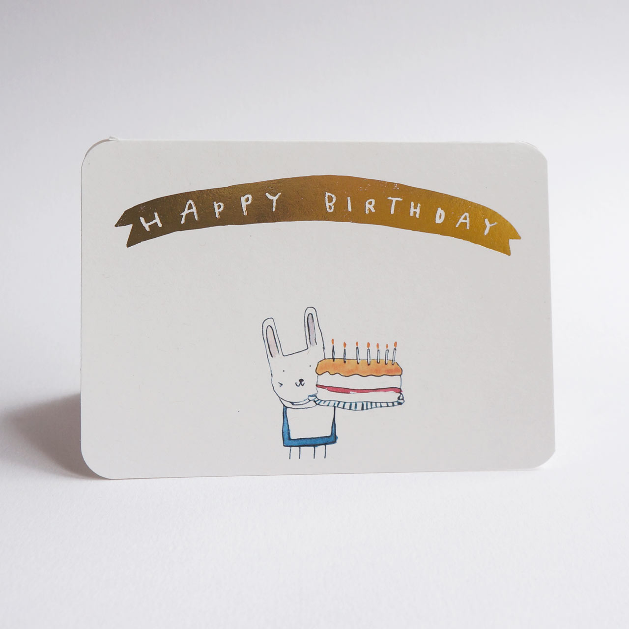 An A6 cream greeting card with rounded corners, featuring a gold banner that reads 'HAPPY BIRTHDAY' above a simple illustration of a white rabbit holding a birthday cake with seven lit candles. 