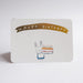 An A6 cream greeting card with rounded corners, featuring a gold banner that reads 'HAPPY BIRTHDAY' above a simple illustration of a white rabbit holding a birthday cake with seven lit candles. 