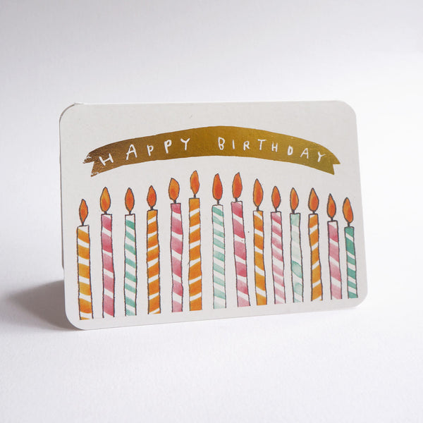 A rectangular birthday card with rounded corners, featuring a gold banner that reads 'HAPPY BIRTHDAY' above a row of colorful striped candles, photographed on a white background.