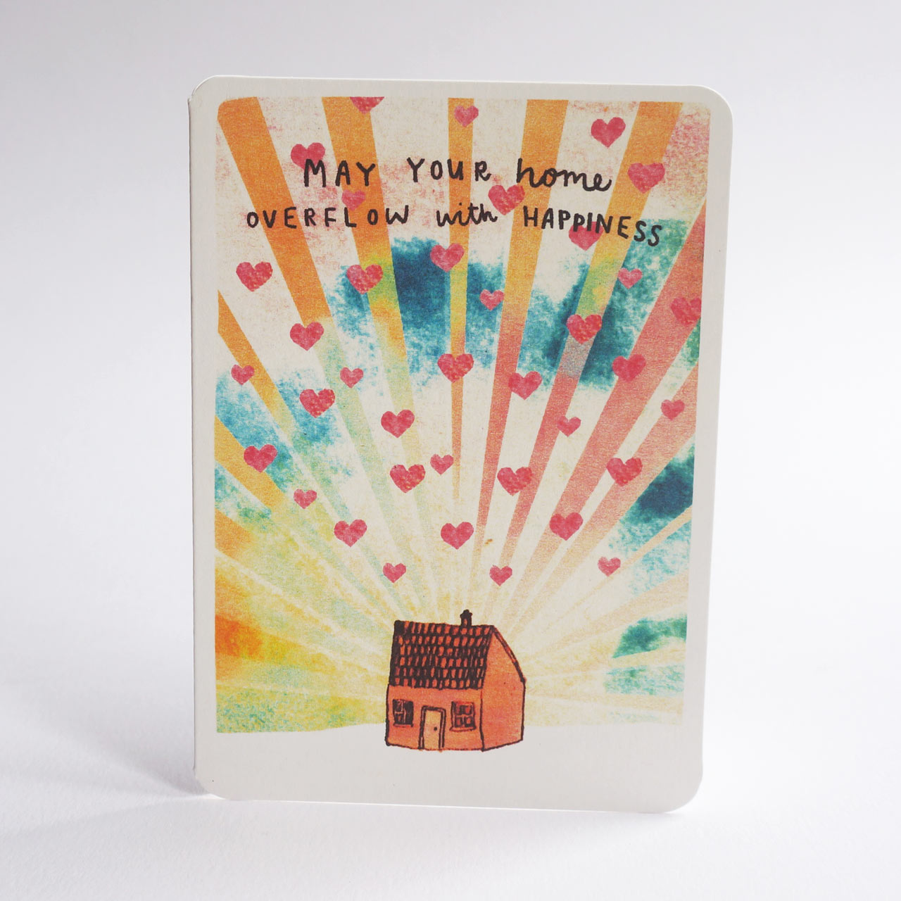 An A6 greeting card with rounded corners, featuring a colorful watercolor-style illustration of sun rays, scattered pink hearts, and a small house at the bottom. Handwritten text at the top reads, 'MAY YOUR HOME OVERFLOW WITH HAPPINESS.