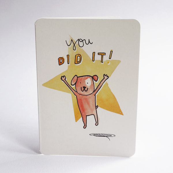 An A6 congratulations card with rounded corners, featuring a hand-drawn illustration of an orange dog jumping in front of a yellow star with the text 'you DID IT!' on a white background.