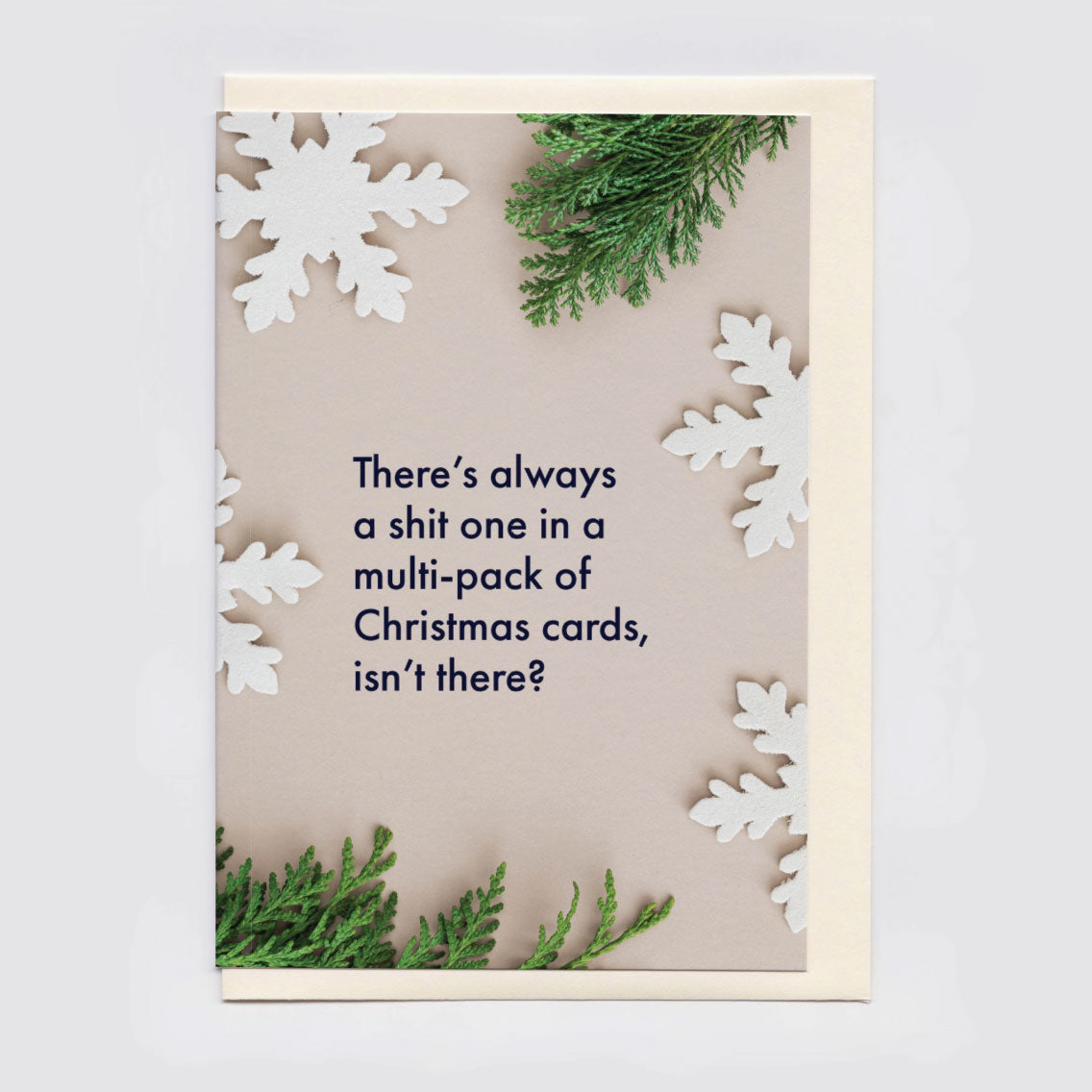 A Christmas greeting card with a beige background, featuring the text 'There's always a shit one in a multi-pack of Christmas cards, isn't there?' in dark navy lettering. The card is decorated with white paper snowflakes and green pine branches, shown with a cream envelope against a light gray background.