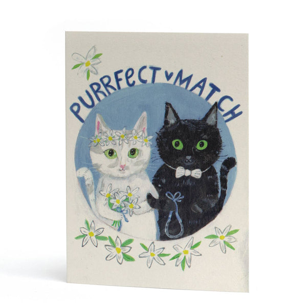 An A6 greeting card featuring an illustration of a white cat and a black cat both with green eyes, linking arms, in their wedding finest. The text above them reads 'PURRFECT MATCH'. The card has a cream-colored background and is shown standing upright against a white studio background.