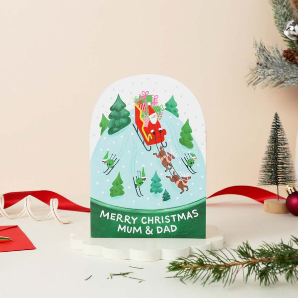 Die-cut snow globe card with Santa in his sleigh pulled by reindeer over snowy Christmas trees. Text reads: Merry Christmas Mum and Dad.
