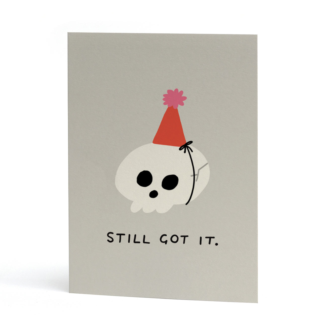 A minimalist A6 card with a light grey background featuring a cartoon skull wearing a red party hat with a pink pom-pom, and the text 'STILL GOT IT.' printed below.
