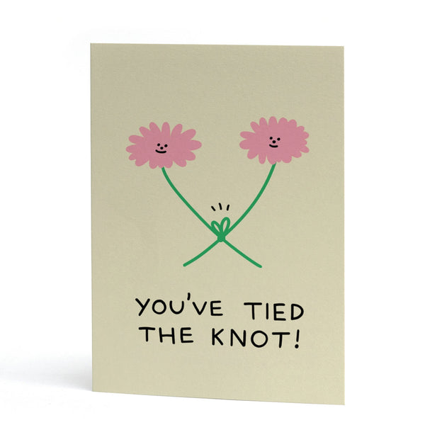 An A6 greeting card with a light beige background, featuring a hand-drawn illustration of two smiling pink flowers with green stems tied together, and the text 'YOU'VE TIED THE KNOT!' in black below.