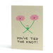 An A6 greeting card with a light beige background, featuring a hand-drawn illustration of two smiling pink flowers with green stems tied together, and the text 'YOU'VE TIED THE KNOT!' in black below.